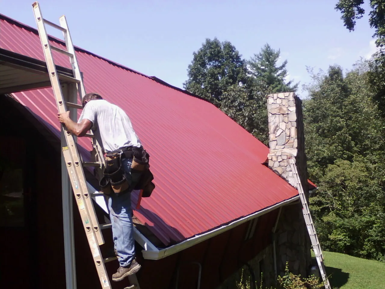 Roofer on a ladder beside a red metal roof and stone chimney — Soffit & Fascia Repair by Roofing Brothers in Mountain Ranch